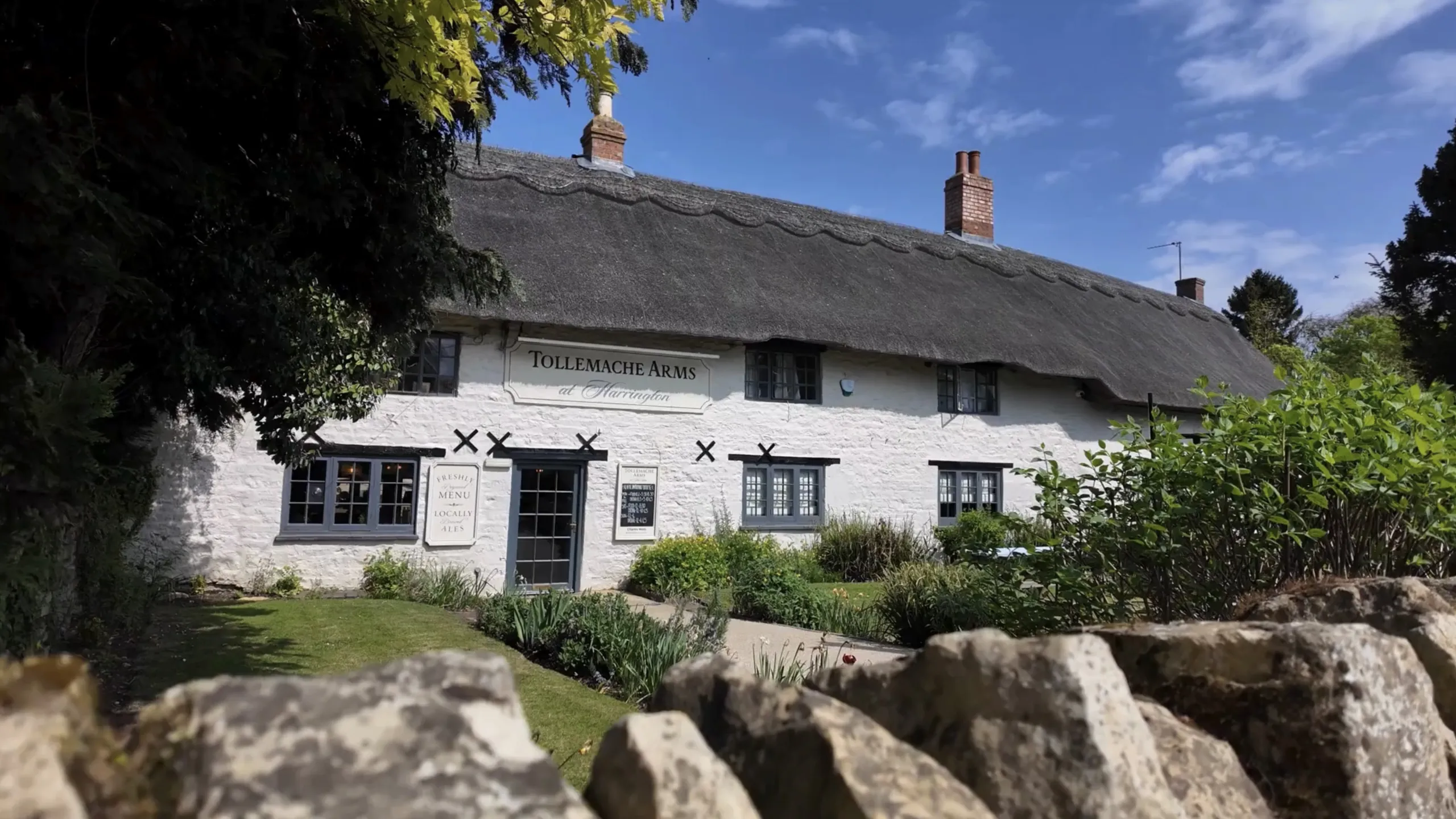 White stone building with a thatched roof and garden in front under a blue sky, named Tollemache Arms at Harrington.