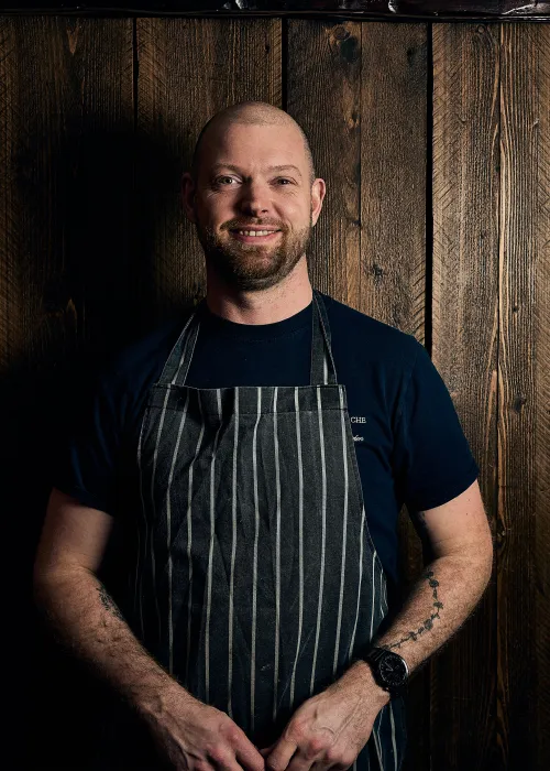 Smiling bald man wearing a striped apron and black t-shirt standing against a wooden plank wall.
