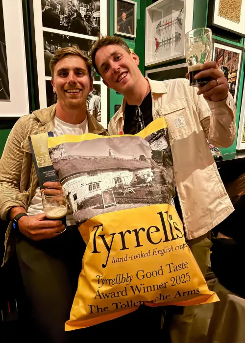 Two smiling men holding drinks and a large Tyrrells crisps bag with award text, posing in a pub with framed photos on green walls.