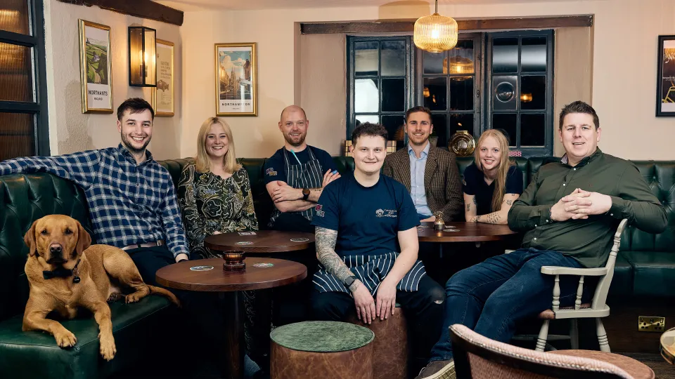 Group of six people and a dog sitting together on green leather sofas in a cozy room with framed pictures and a large window behind them.