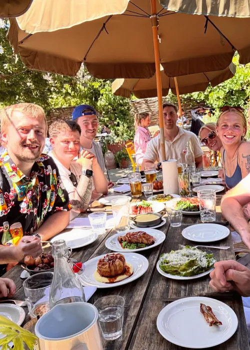 Group of people smiling and sitting around a wooden table outdoors with plates of food and drinks under beige umbrellas.