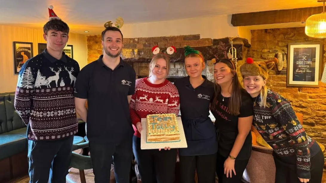 Group of six people indoors, some wearing festive hats and sweaters, with one person holding a cake decorated with icing words.