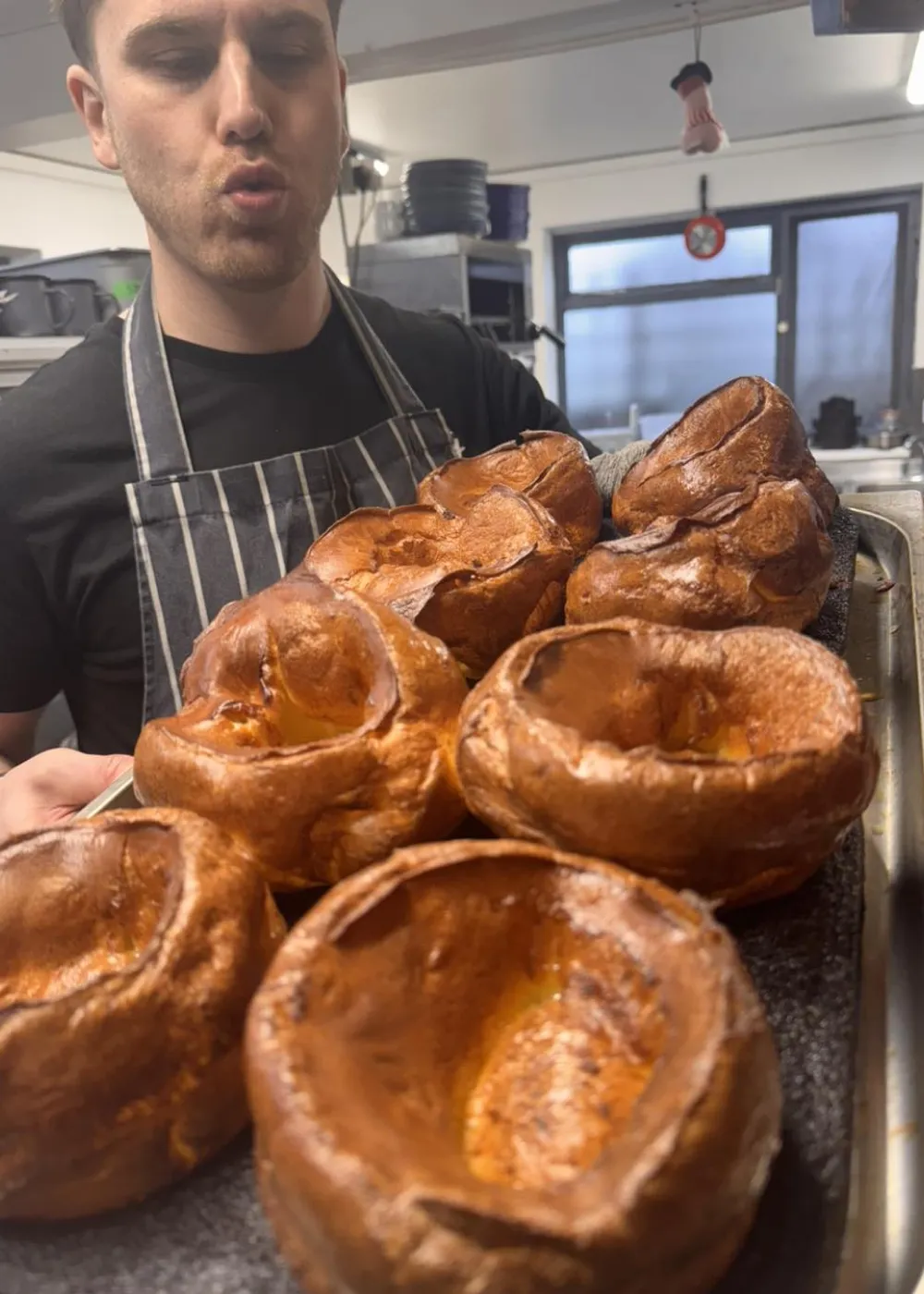 Man in a striped apron holding a tray with freshly baked Yorkshire puddings in a kitchen.