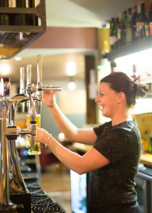 Woman bartender smiling while pouring a draft beer from a tap in a well-lit bar.
