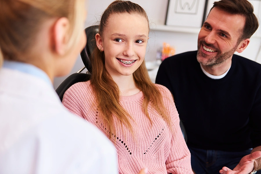 Smiling teenage girl with braces talking to a female dentist while a man, possibly her father, looks on and smiles.