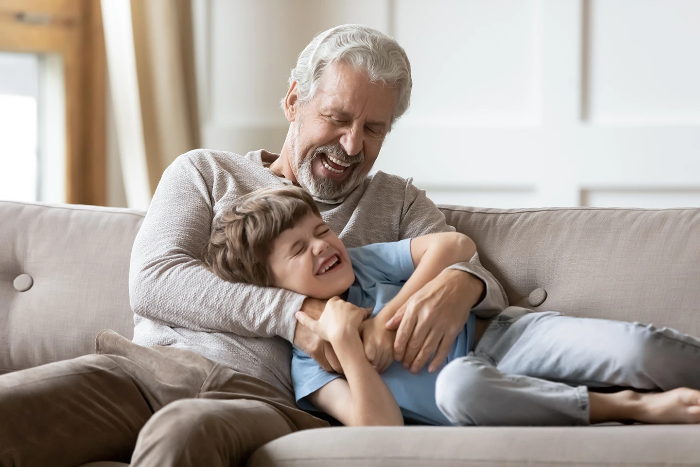 Smiling older man tickling a laughing young boy lying on a beige couch.