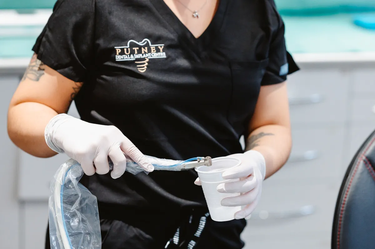 Dental professional in black uniform and gloves holding a dental tool and a white cup in a clinic setting.