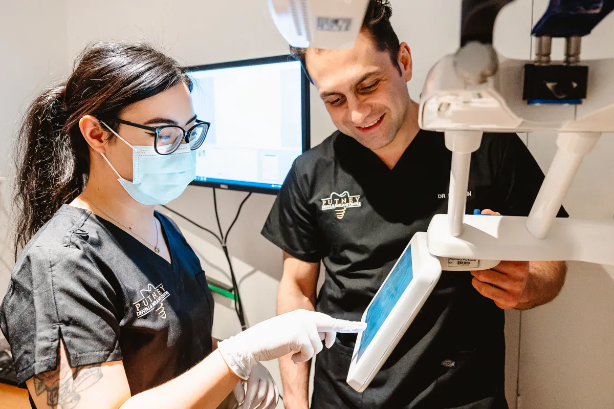 Two dental professionals in black scrubs operate a touchscreen device in a dental office.