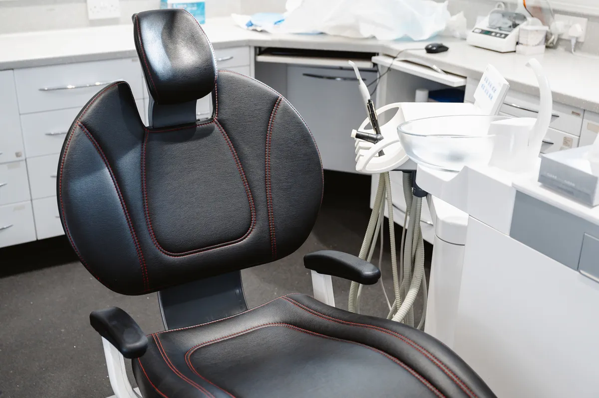 Black leather dental chair with red stitching in a dental clinic, surrounded by medical equipment and white cabinetry.