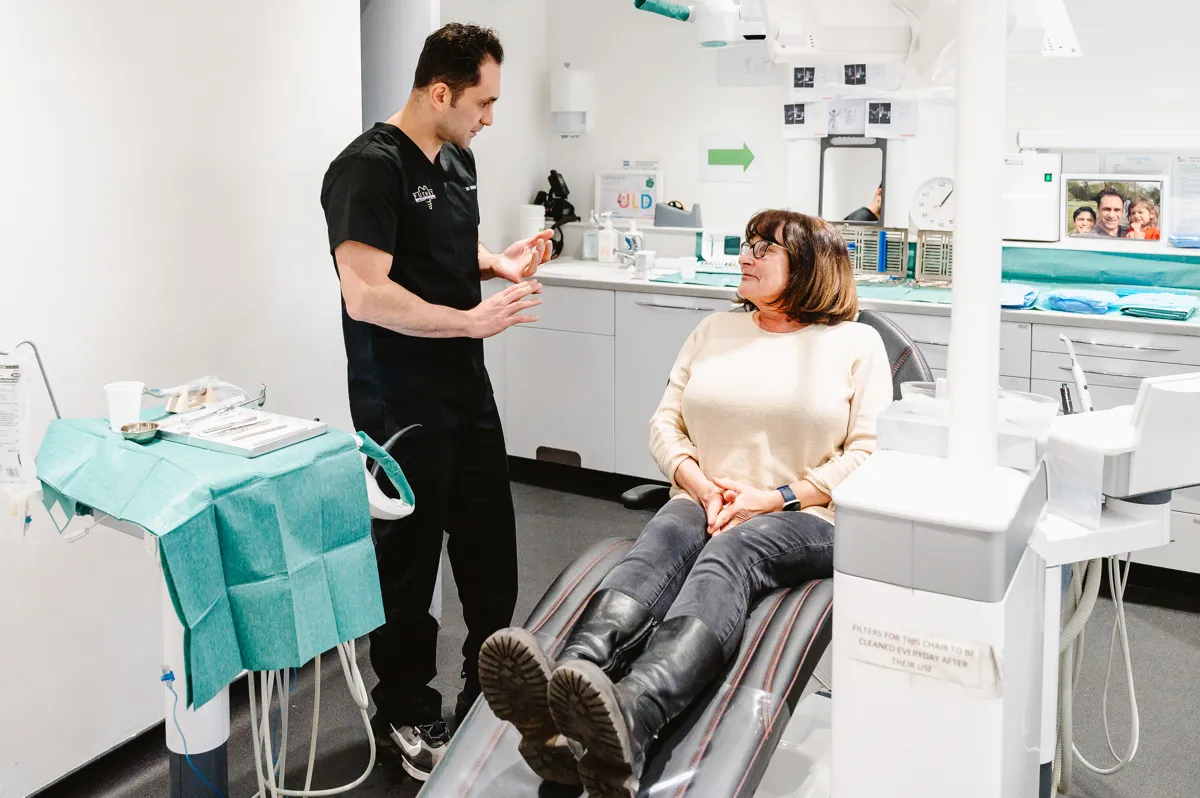 Dentist in black scrubs explaining a procedure to a seated female patient in a dental clinic.