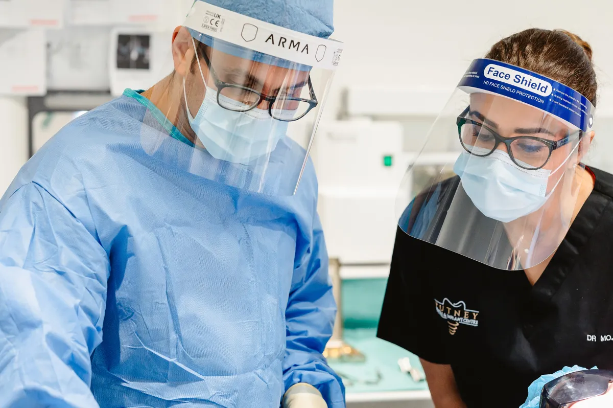 Two medical professionals wearing face shields, masks, and glasses, focusing on a task in a clinical setting.