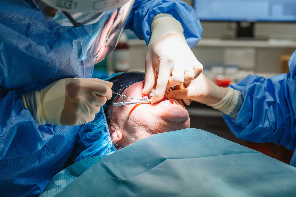 Dentist in blue protective gown and gloves performing a dental procedure on a patient lying down with mouth open.