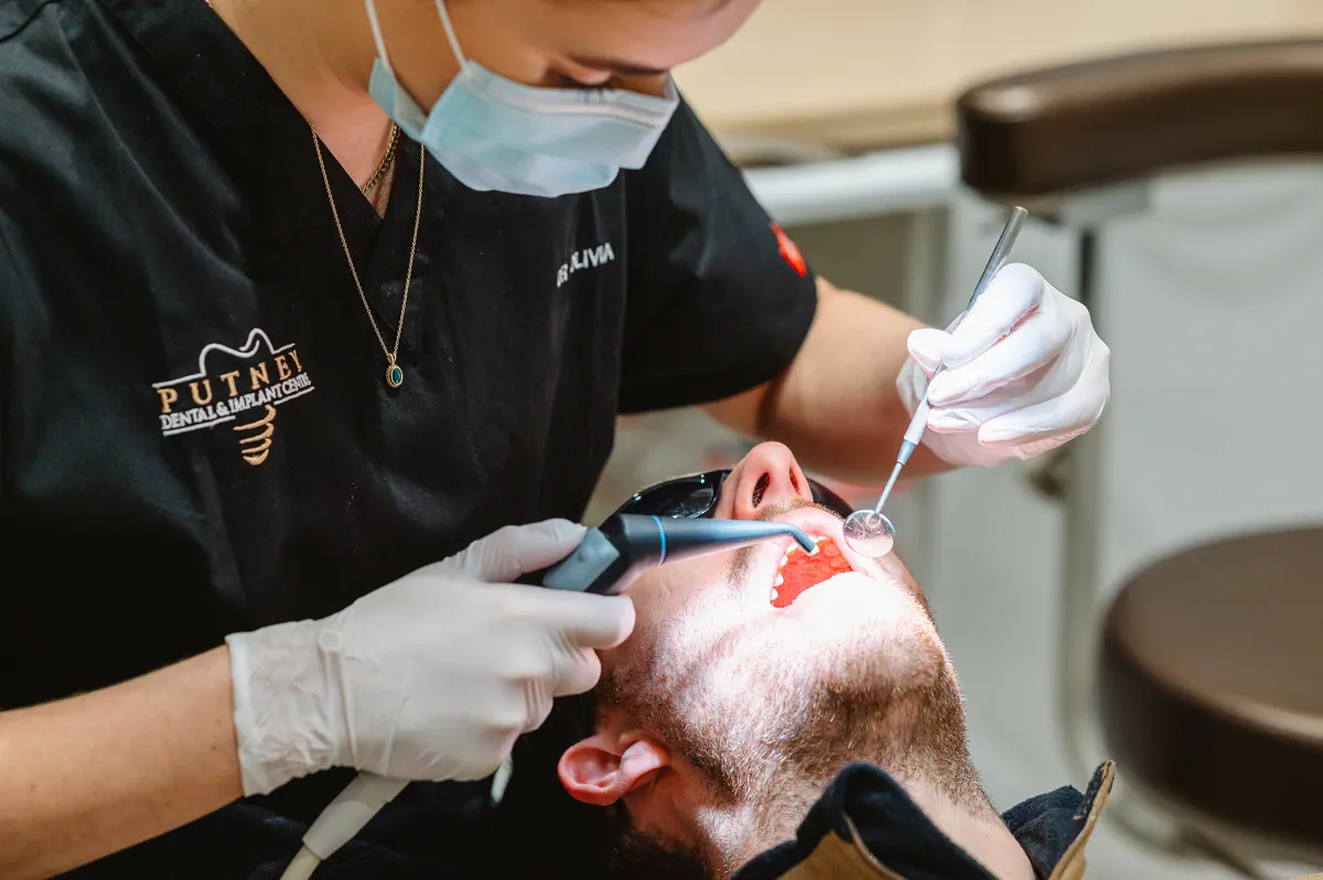 Dentist wearing a mask and gloves performing a dental checkup on a male patient using dental instruments.