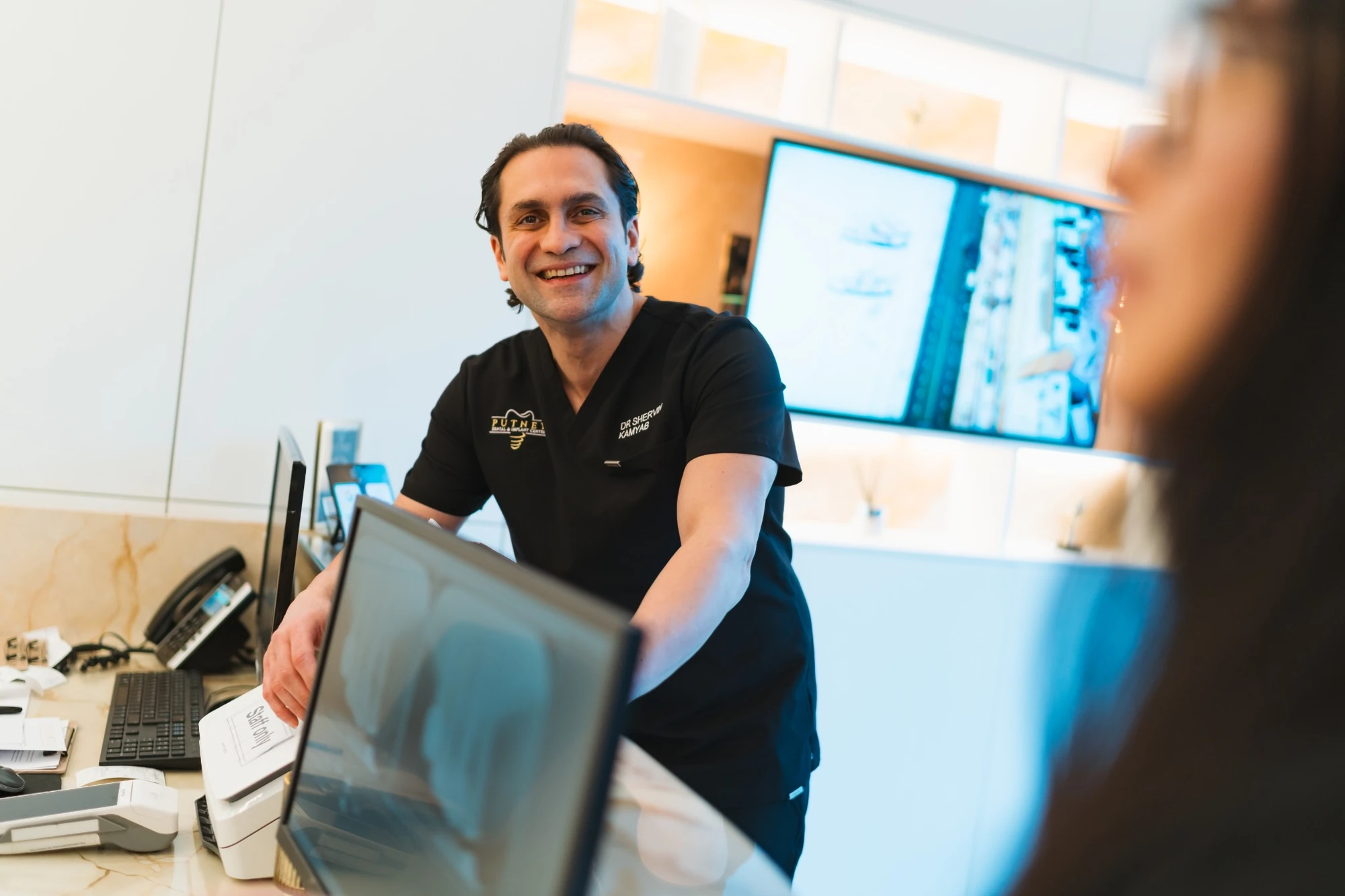 Smiling male dentist in black scrubs standing behind a reception desk, talking to a patient.