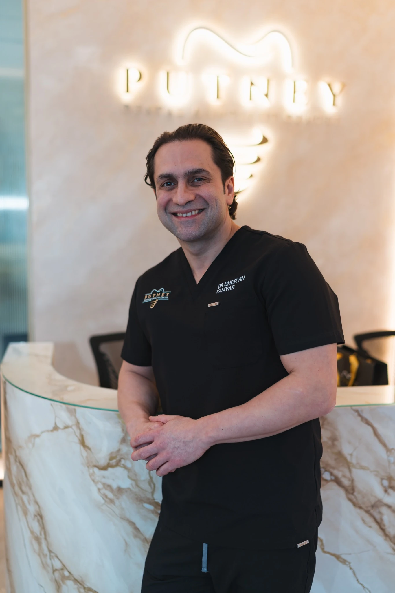 Smiling man in black medical scrubs standing in front of a marble reception desk with a glowing 'PUTNEY' sign behind him.