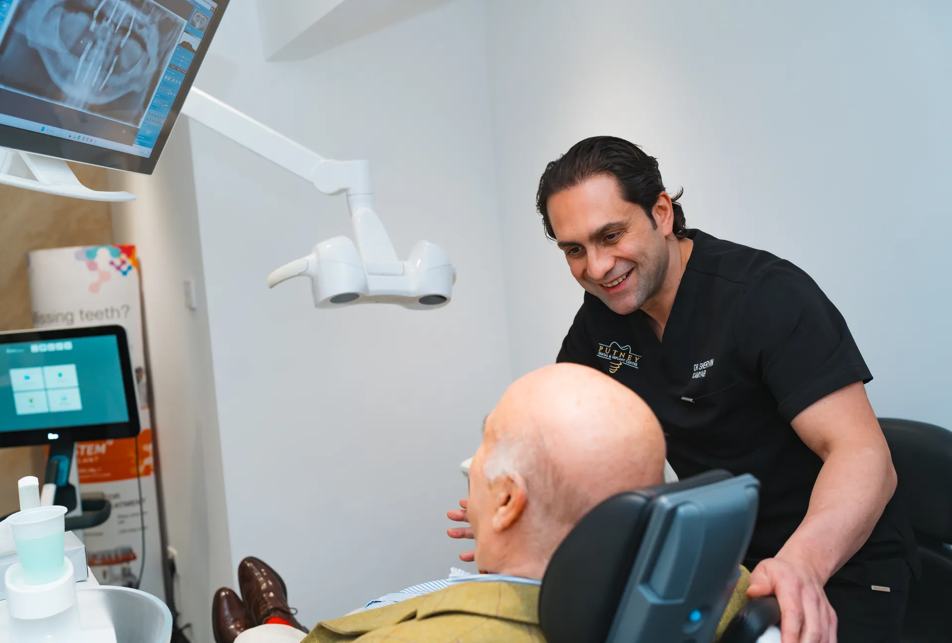 Dentist smiling and talking to an elderly male patient in a dental chair with a dental X-ray displayed on a monitor.