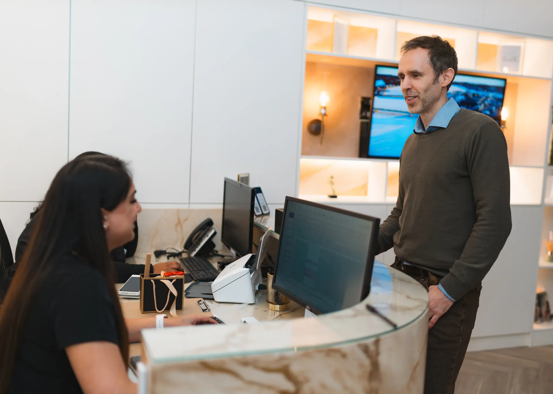 Man standing and talking to a smiling woman sitting behind a curved marble reception desk with computer monitors.