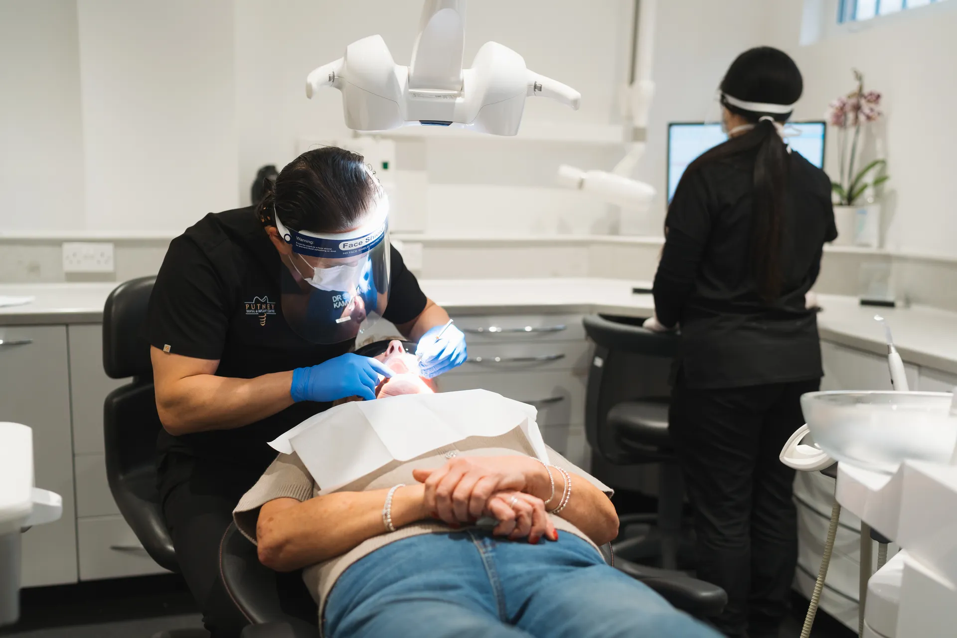 Dentist wearing face shield and gloves examining a patient's mouth in a dental clinic while an assistant works on a computer in the background.