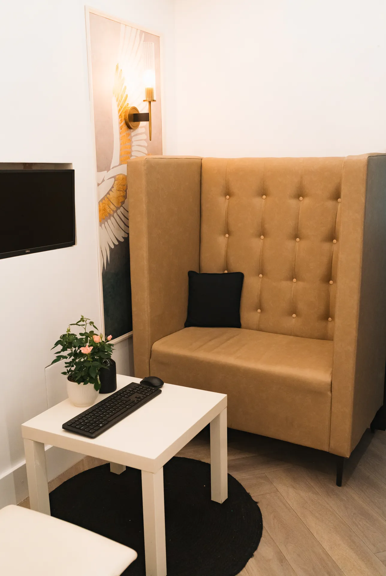 Cozy corner with a tan tufted high-back sofa, white table holding a black keyboard, mouse, and a potted plant with pink flowers, and a wall-mounted flat screen TV.