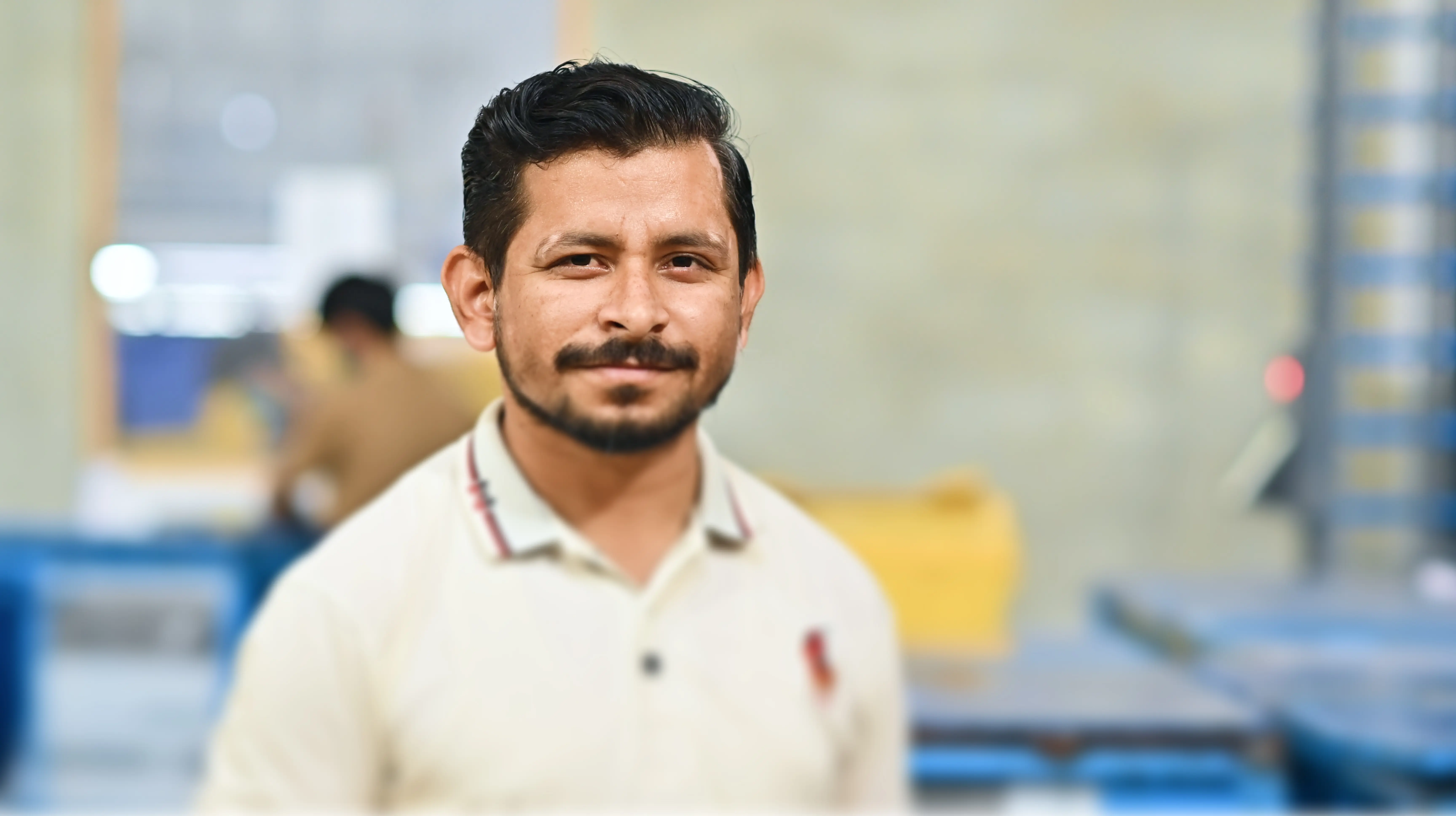 Man with short dark hair and beard wearing a light-colored polo shirt, standing indoors with blurred industrial background.