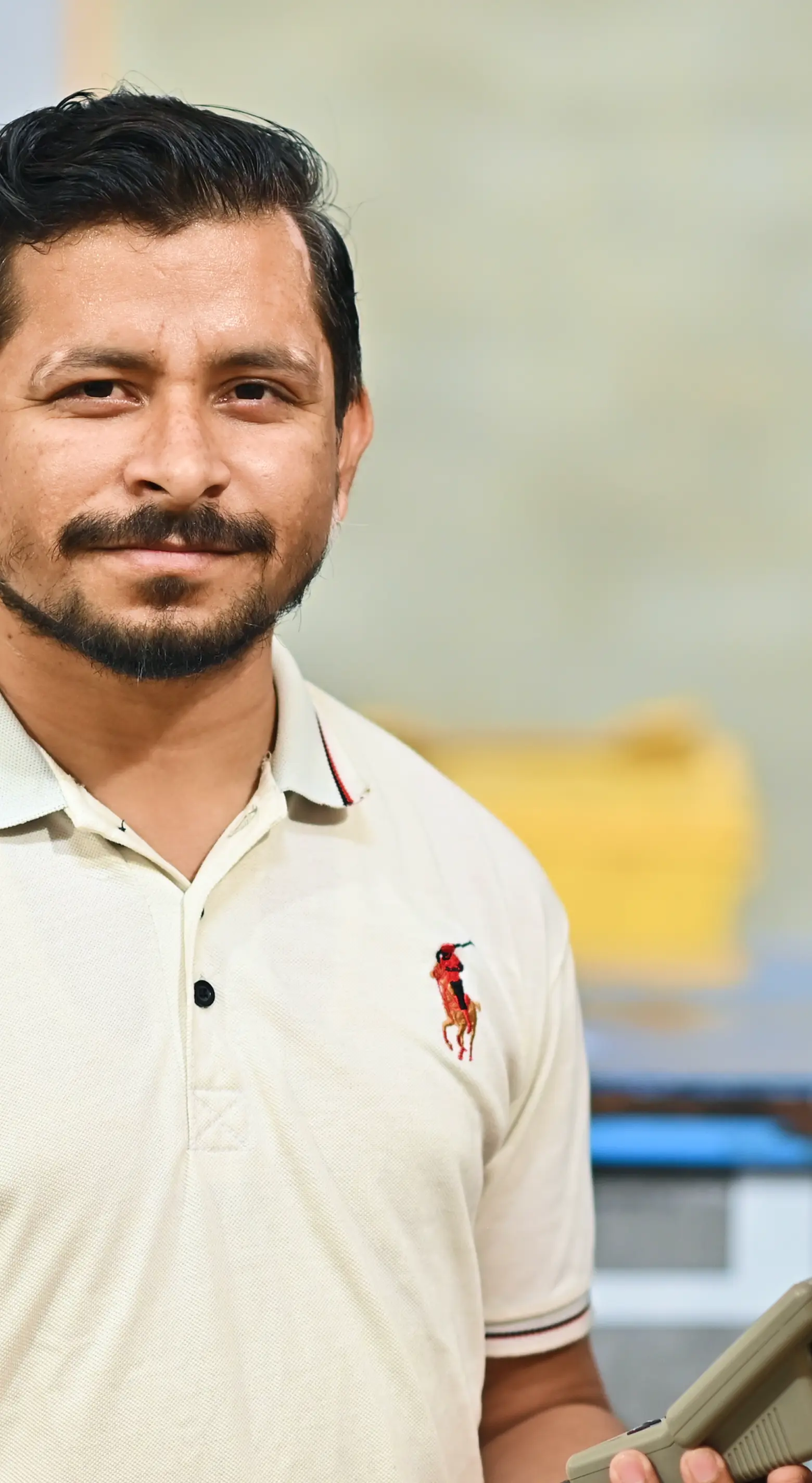 Smiling man with a beard wearing a white polo shirt holding a handheld device.
