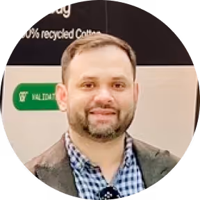 Smiling man with short hair and beard wearing a checkered shirt and dark jacket in a circular frame.