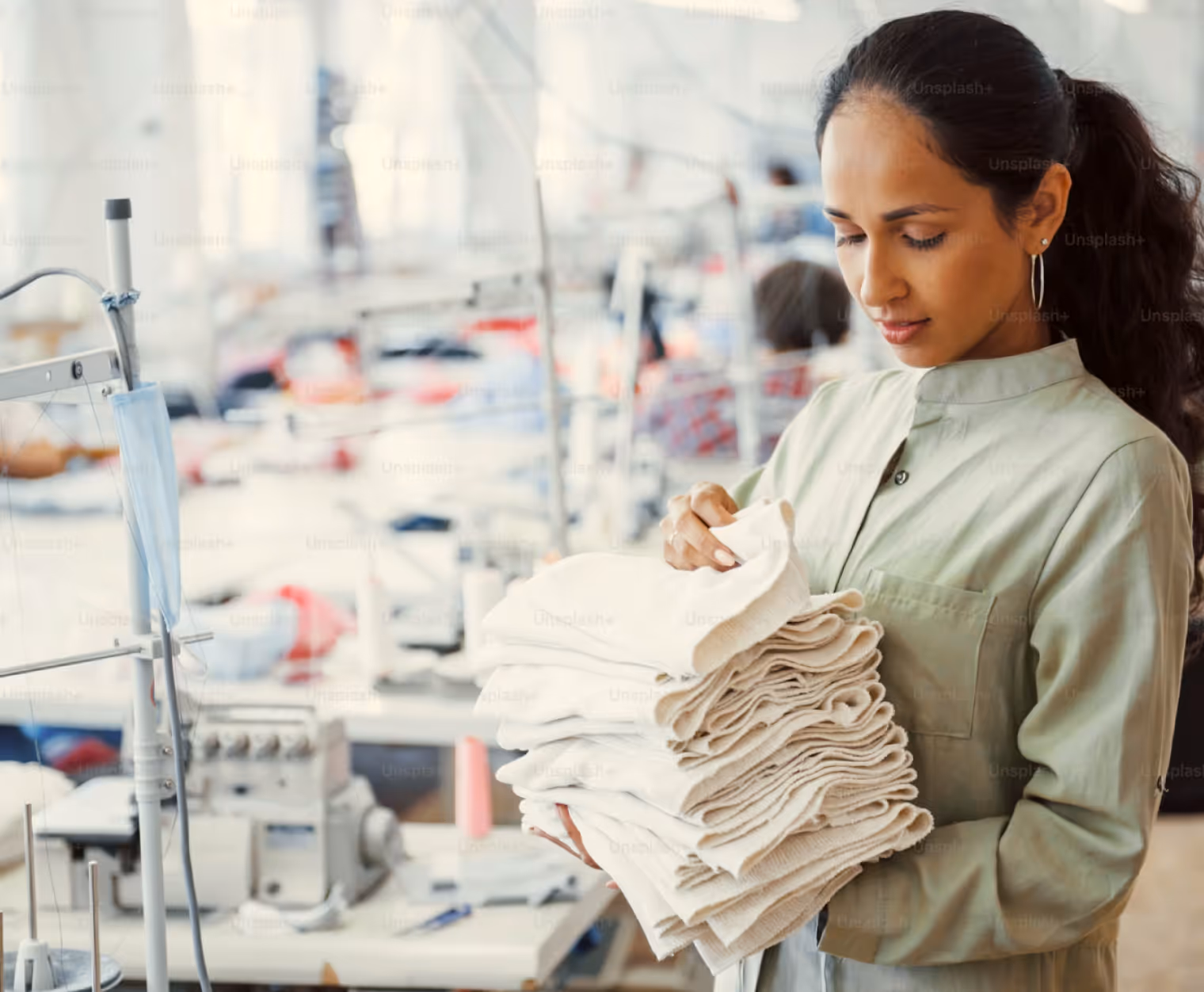 Woman in a factory holding and inspecting folded white fabric pieces, with sewing machines in the background.