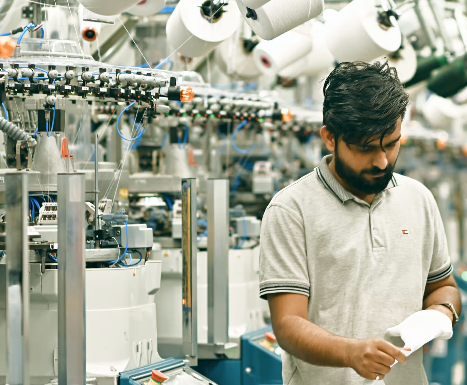 Man in a factory inspecting a white textile product amid industrial knitting machines with spools of yarn above.
