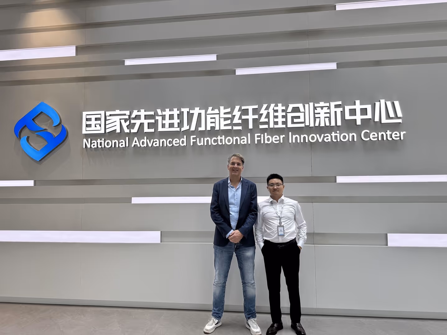 Two men standing in front of a wall with a blue logo and the text National Advanced Functional Fiber Innovation Center in Chinese and English.