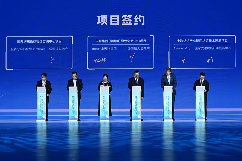 Six people standing on a stage signing agreements under a blue backdrop with three contract sections highlighted above them.