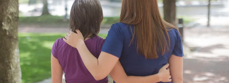 Two people with arms around each other walking outdoors in a park, viewed from behind.