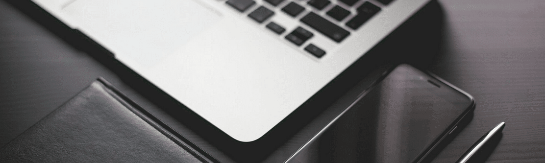 Close-up of a laptop, smartphone, pen, and black notebook on a dark wooden desk.
