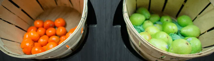 Basket of oranges and basket of green apples on a black wooden surface.