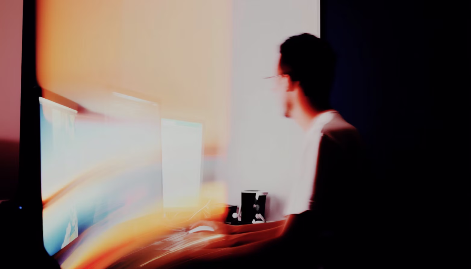 Blurred image of a person working on a computer with bright screen light in a dark room.