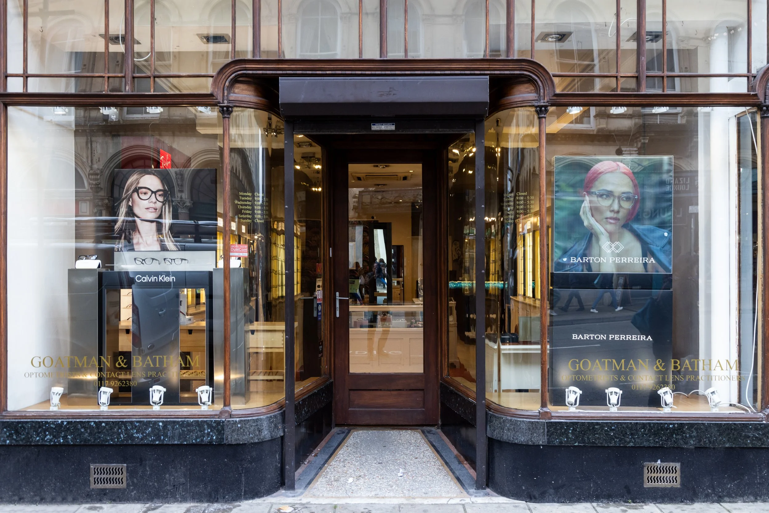 Entrance of Goatman & Batham optometrist store with glass windows displaying Calvin Klein and Barton Perreira eyewear advertisements.