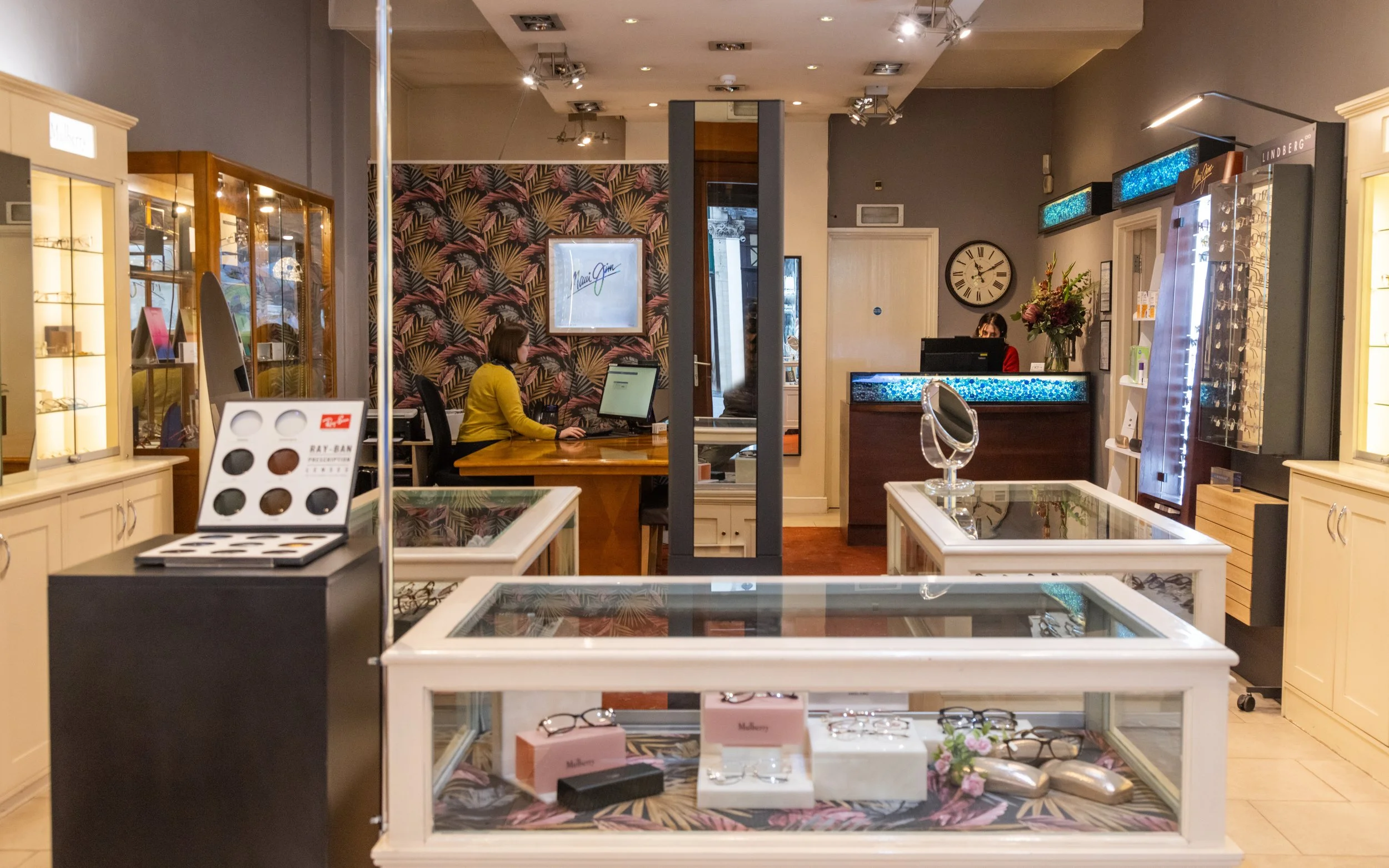 Optical store interior with glass display cases featuring eyeglasses, two employees working at desks, and branded eyewear displays.