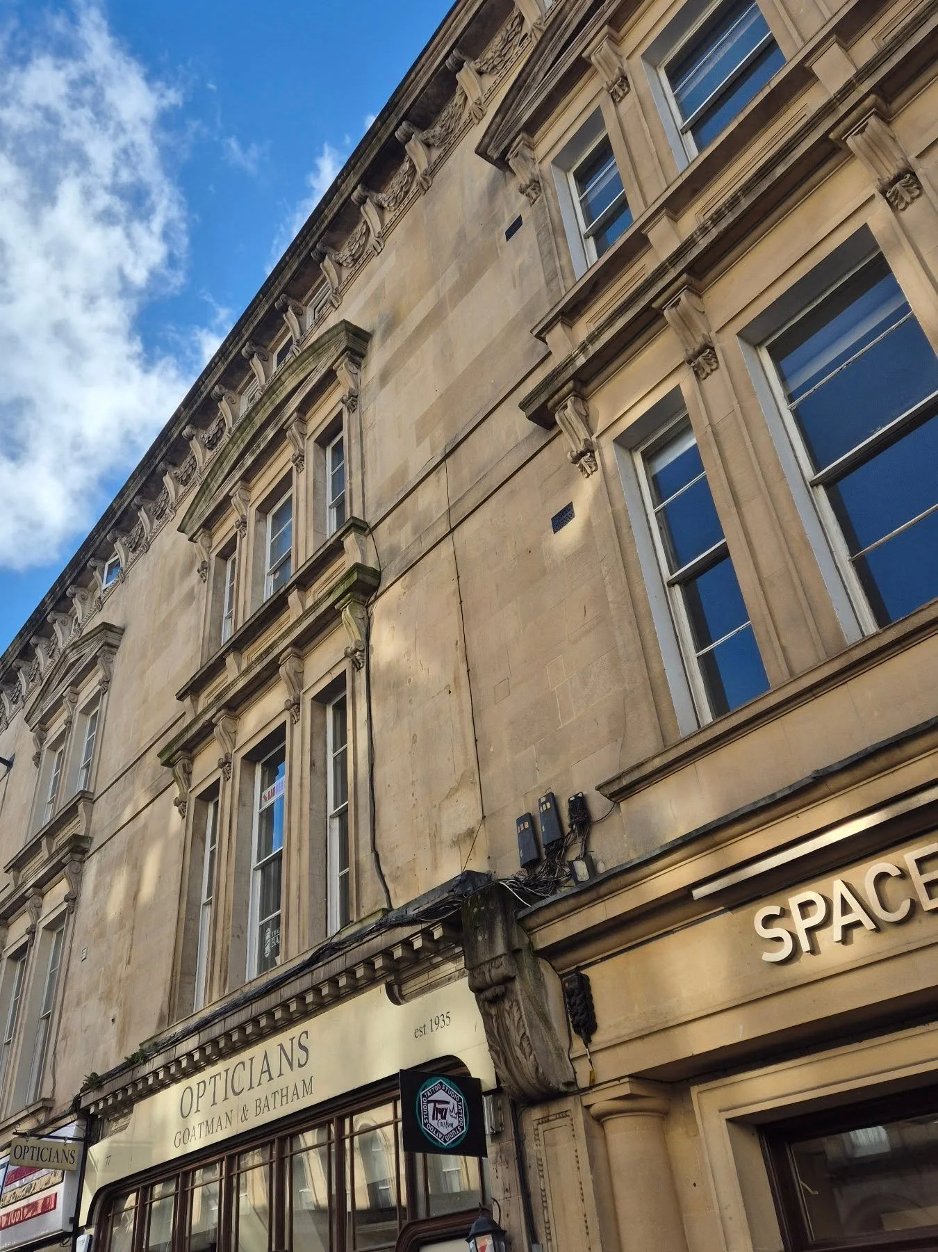 Historic beige stone building with large windows and a sign for Opticians Goatman & Batham established 1935 under a blue sky with some clouds.