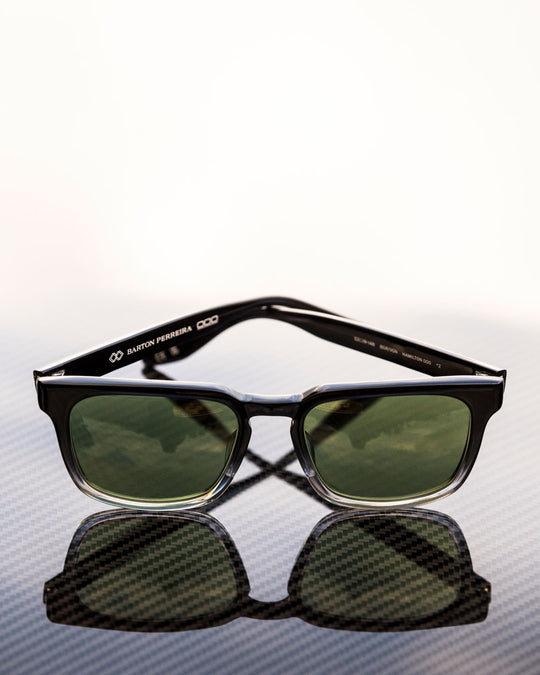 Black-framed Barton Perreira sunglasses with green lenses resting on a reflective carbon fiber surface.