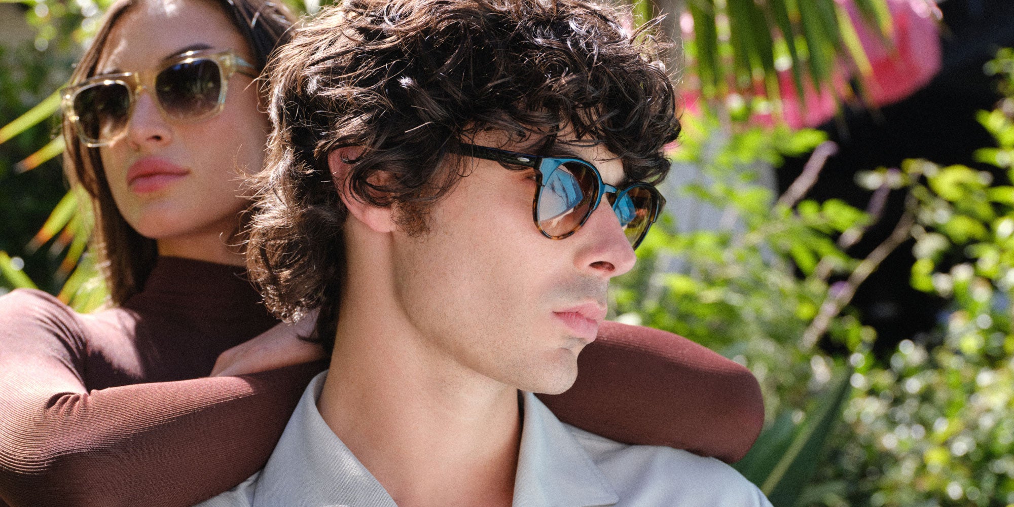 Close-up of a man and woman wearing sunglasses outdoors with green foliage in the background.