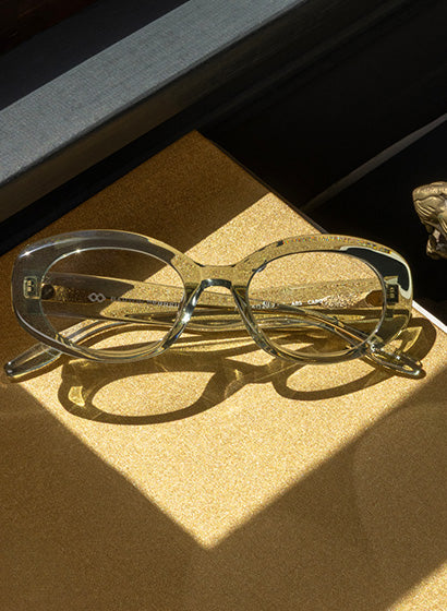 Transparent light green eyeglass frames resting on a golden surface with sunlight casting shadows.