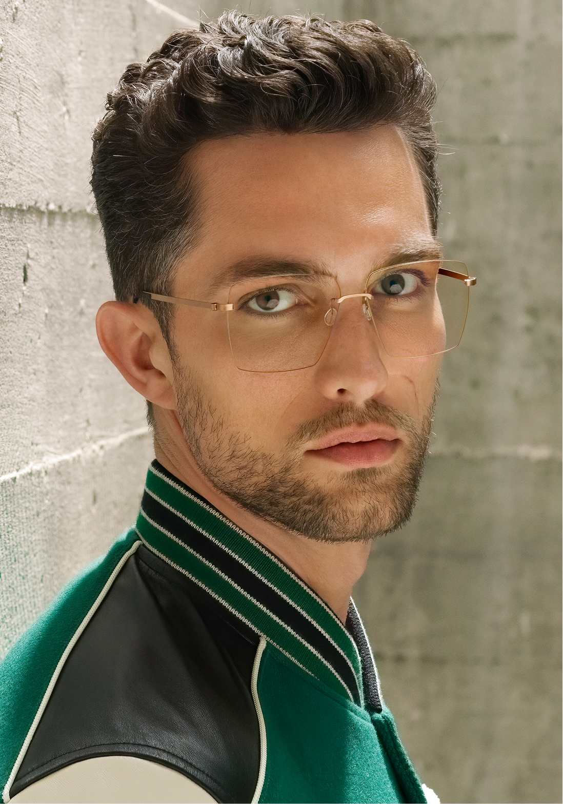 Close-up of a man with curly brown hair and beard wearing clear-framed glasses and a green and black jacket looking at the camera.