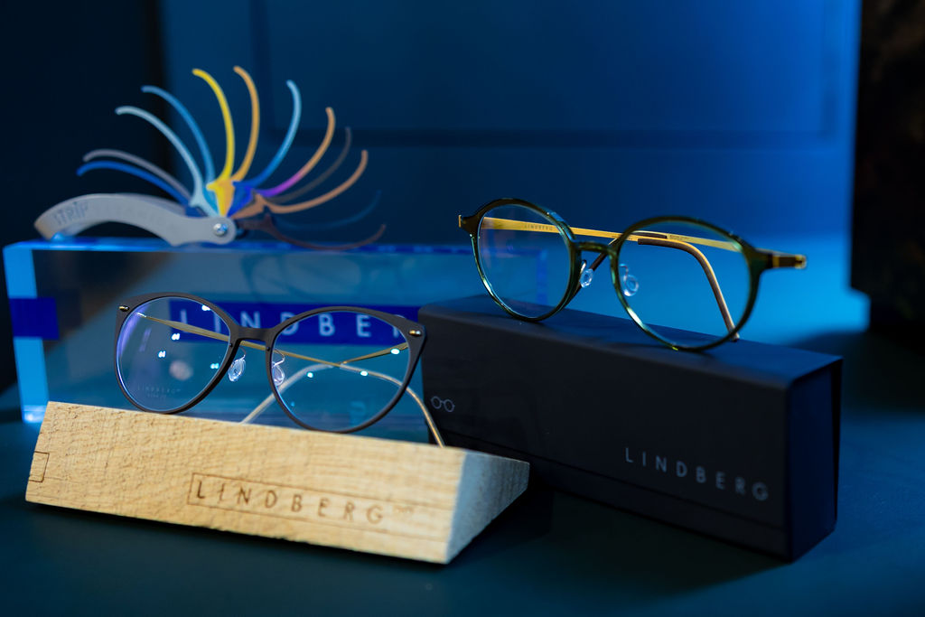 Two pairs of Lindberg eyeglasses displayed on wooden and black holders with a color palette in the background.