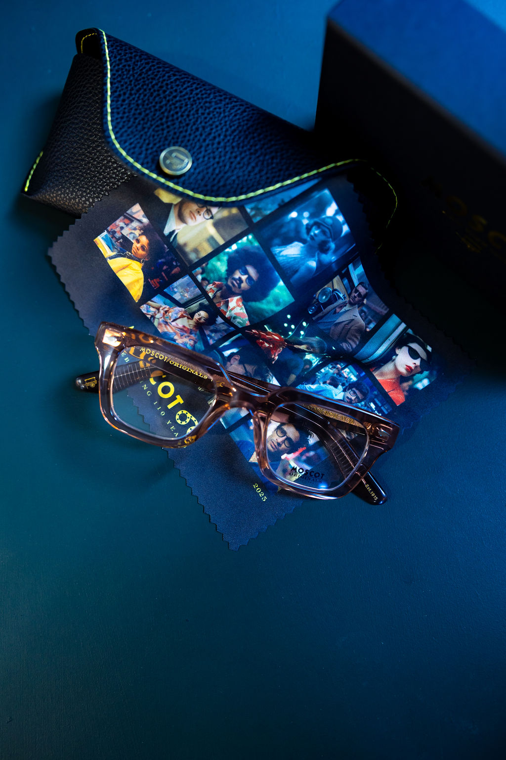 Tortoiseshell eyeglasses resting on a colorful cleaning cloth with vintage portraits, next to a black leather glasses case.