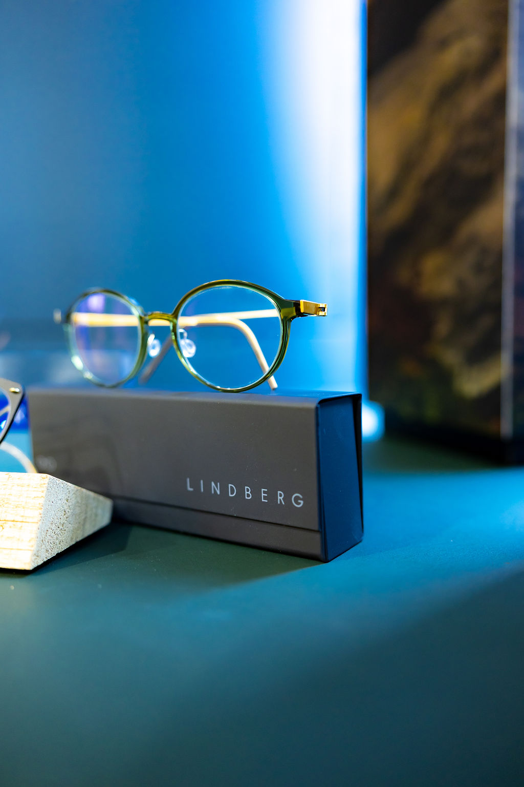 Round eyeglasses with green frames resting on a black Lindberg branded case on a blue surface.