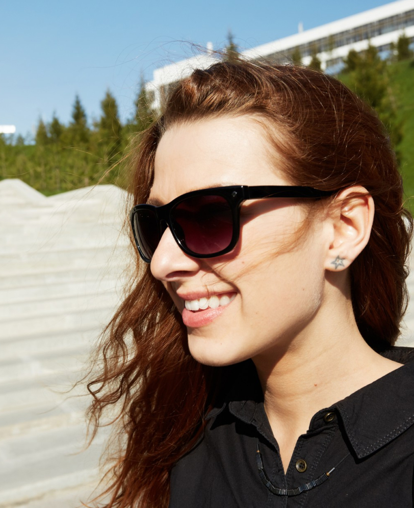 Smiling woman with long red hair wearing black sunglasses, star-shaped earrings, and a black shirt outdoors.