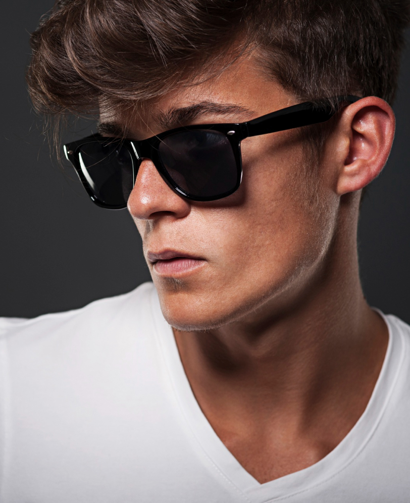 Close-up of a young man with styled brown hair wearing black sunglasses and a white V-neck shirt.