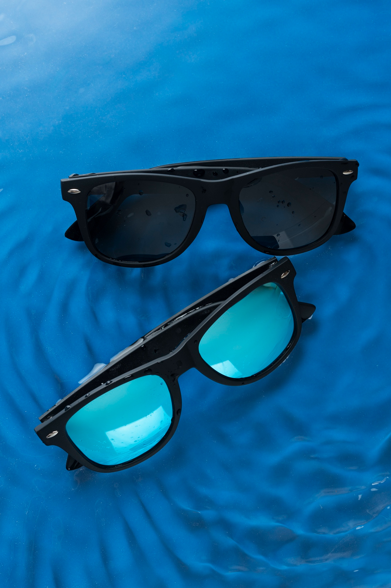 Two pairs of black-framed sunglasses, one with dark lenses and one with blue-tinted lenses, resting on rippling blue water.
