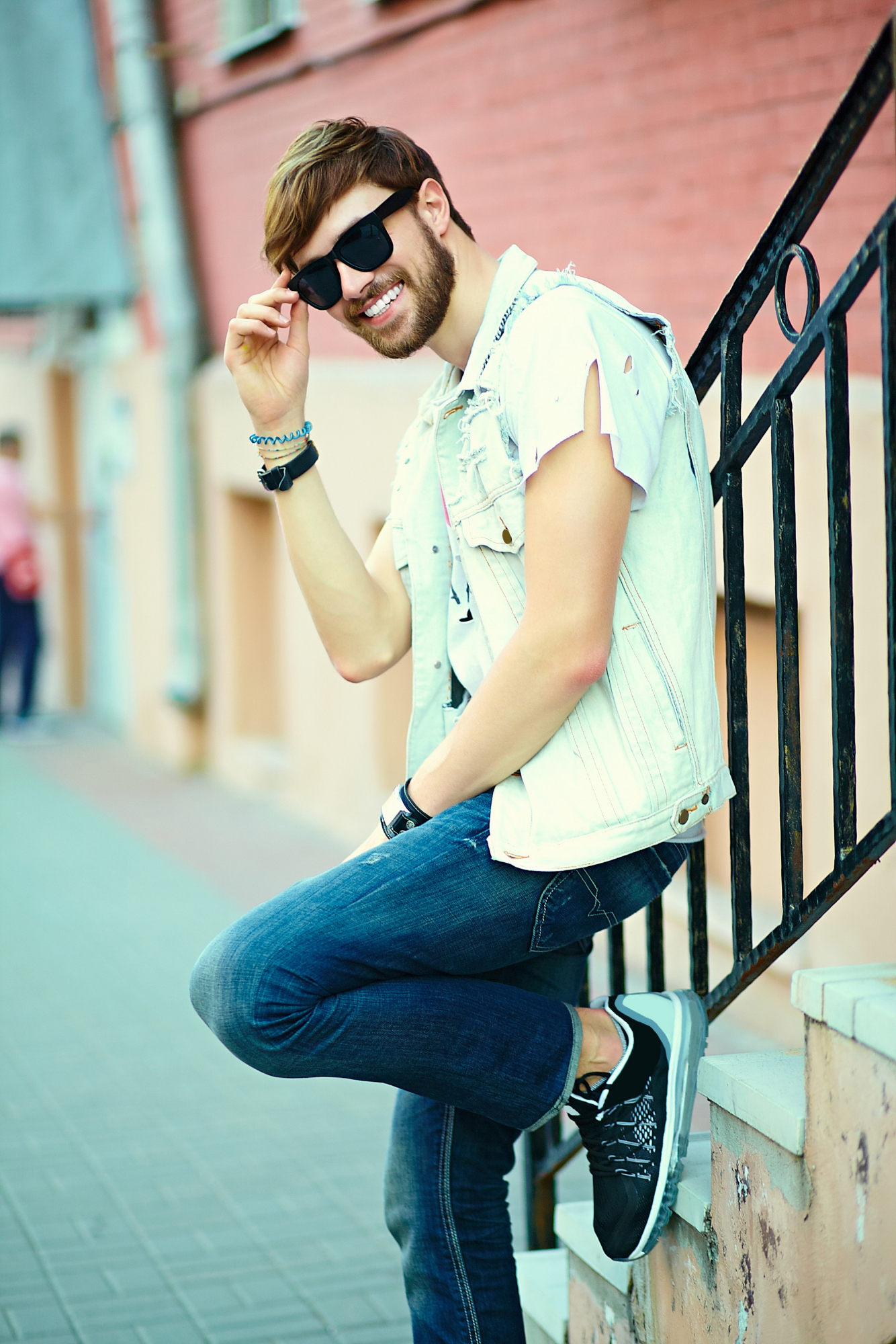 Smiling man wearing sunglasses, a sleeveless denim jacket, and jeans, leaning on a railing on an outdoor staircase.