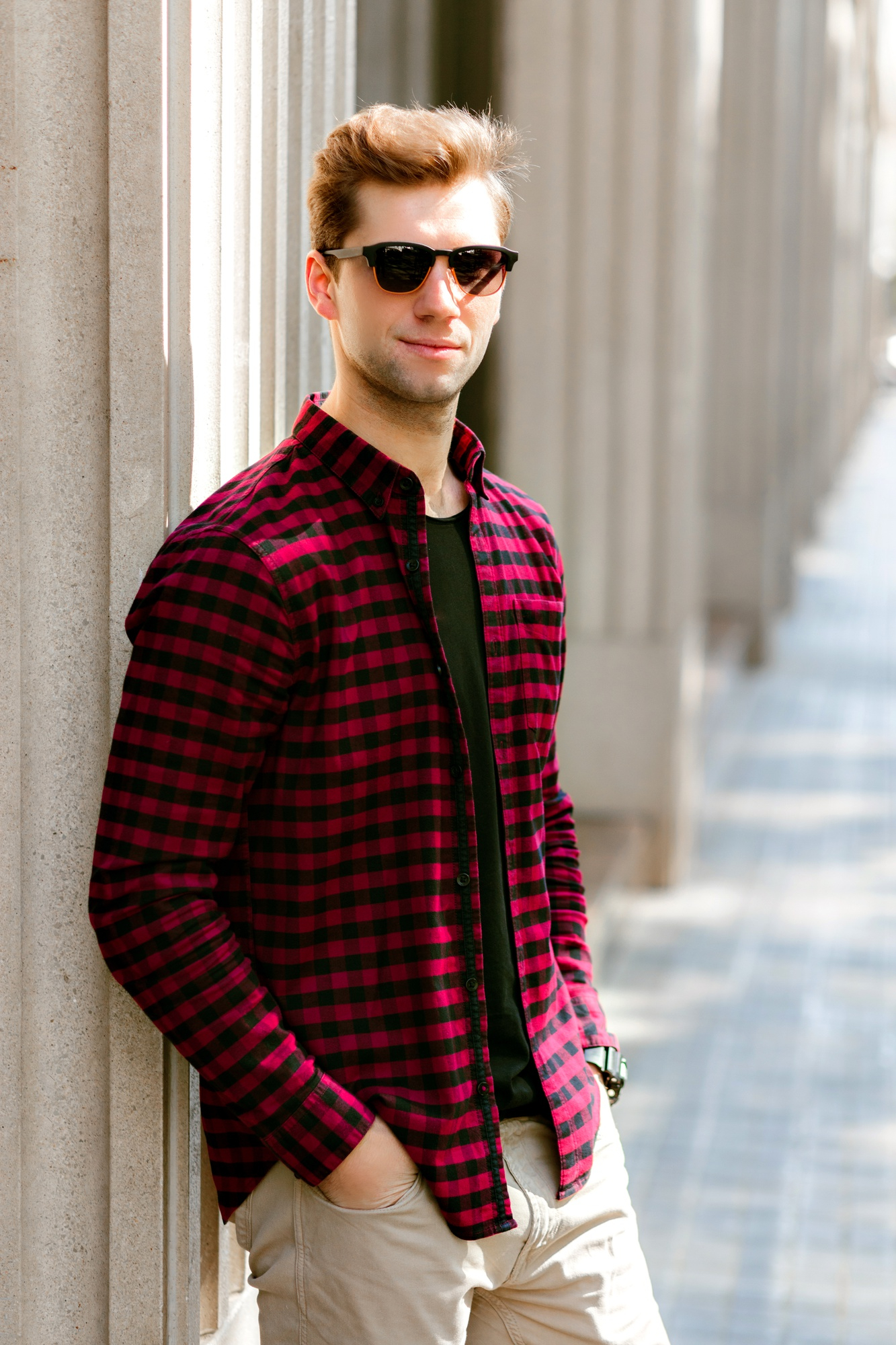 Young man wearing sunglasses, a red and black checkered shirt, and beige pants leaning against a stone column in a sunlit outdoor walkway.