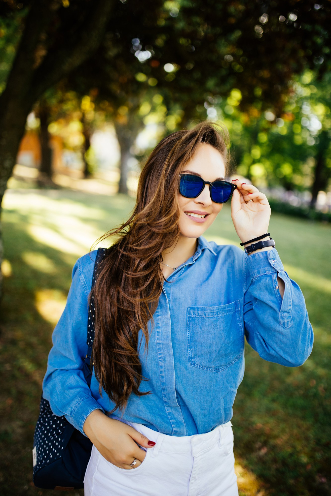 Smiling woman in blue denim shirt and white pants adjusting blue sunglasses outdoors in a park.